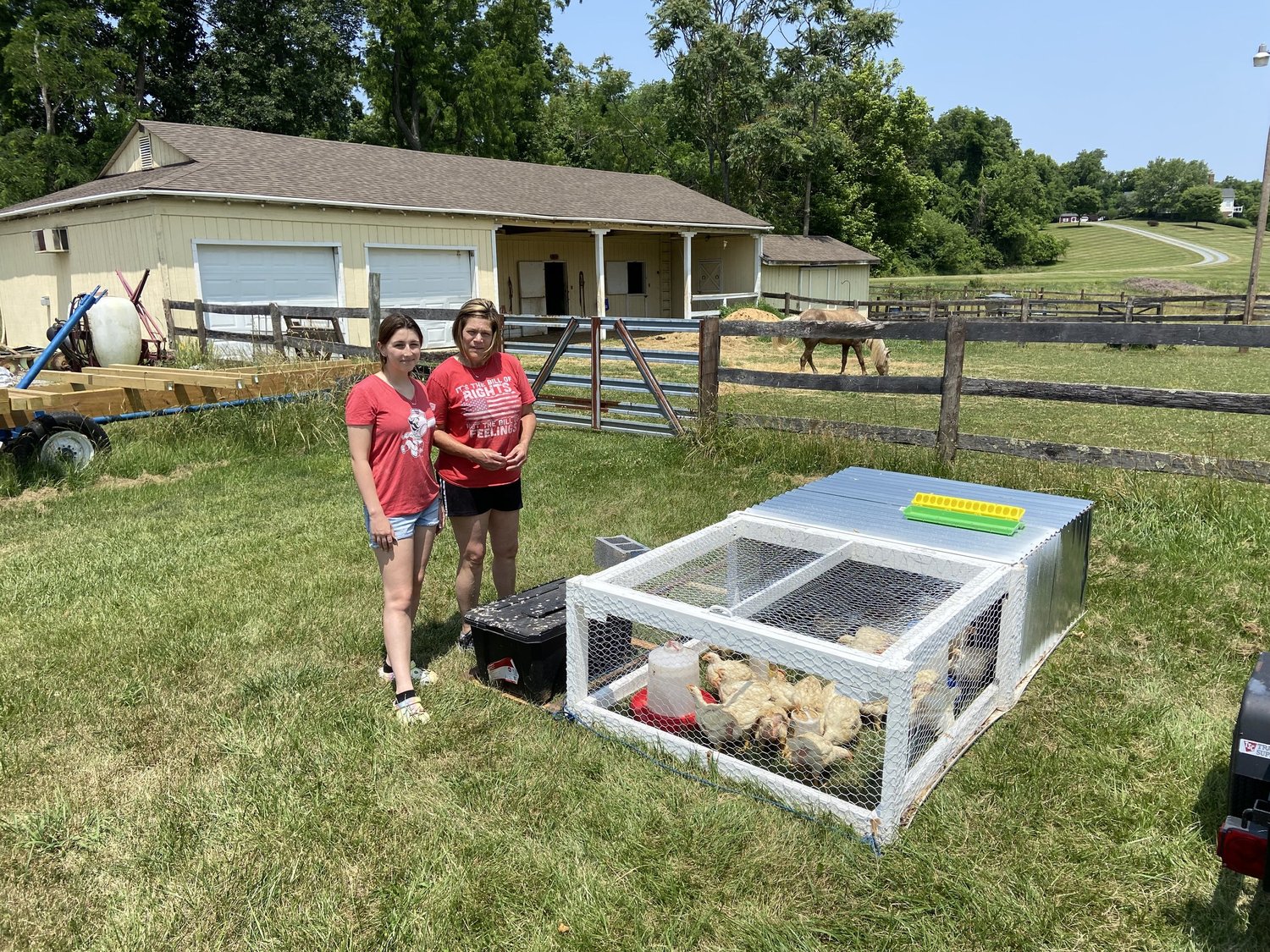 Two women stand next to a chicken coop in a grassy area, with a horse grazing nearby and a barn in the background. The coop is designed for raising chickens and features a metal roof and wire mesh. The scene is set on a sunny day, showcasing a rural farm environment.