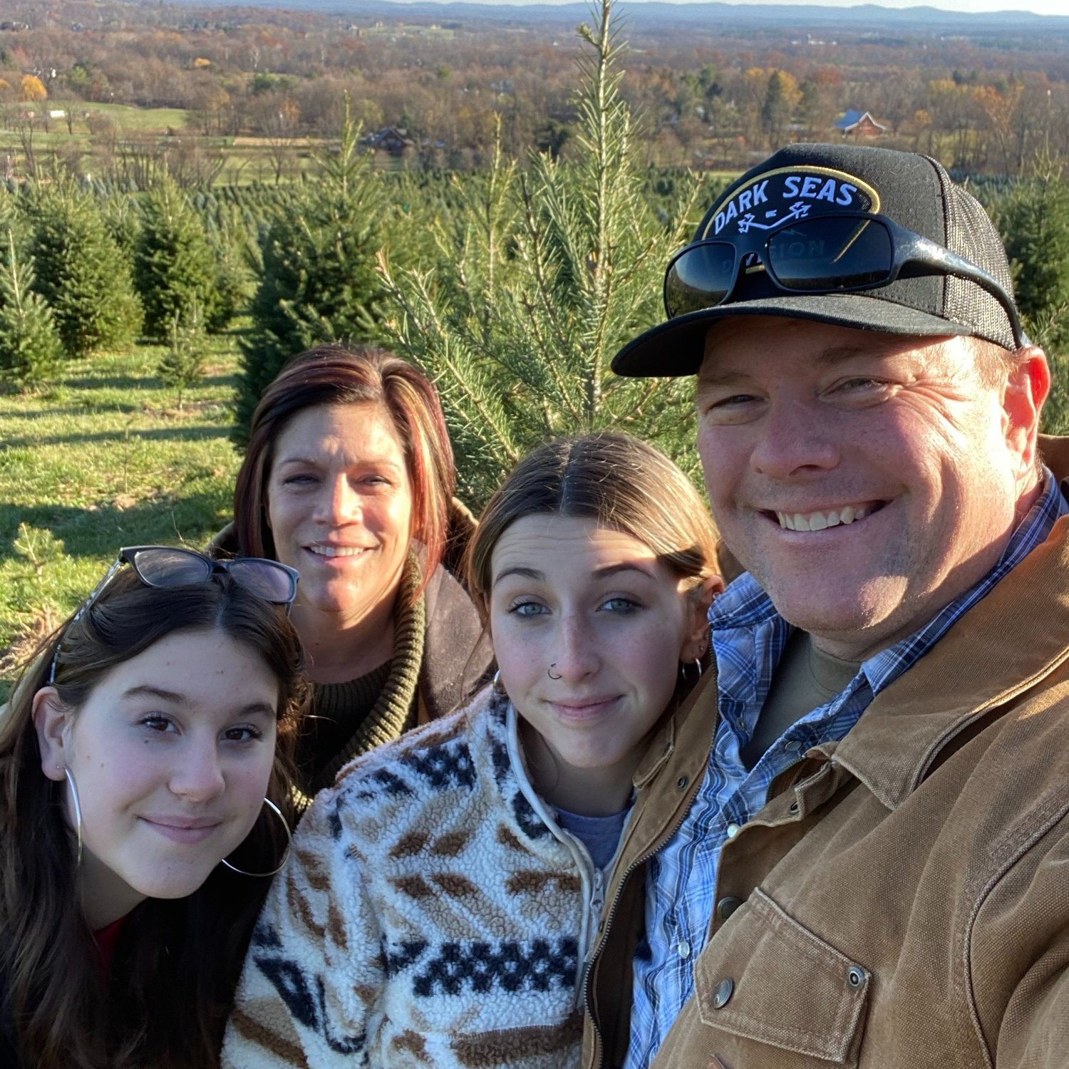 Family enjoying a day outdoors at a Christmas tree farm, surrounded by lush greenery and scenic hills in the background. The group consists of two adults and two teenagers, all smiling and dressed in cozy, casual attire, capturing a joyful holiday tradition.