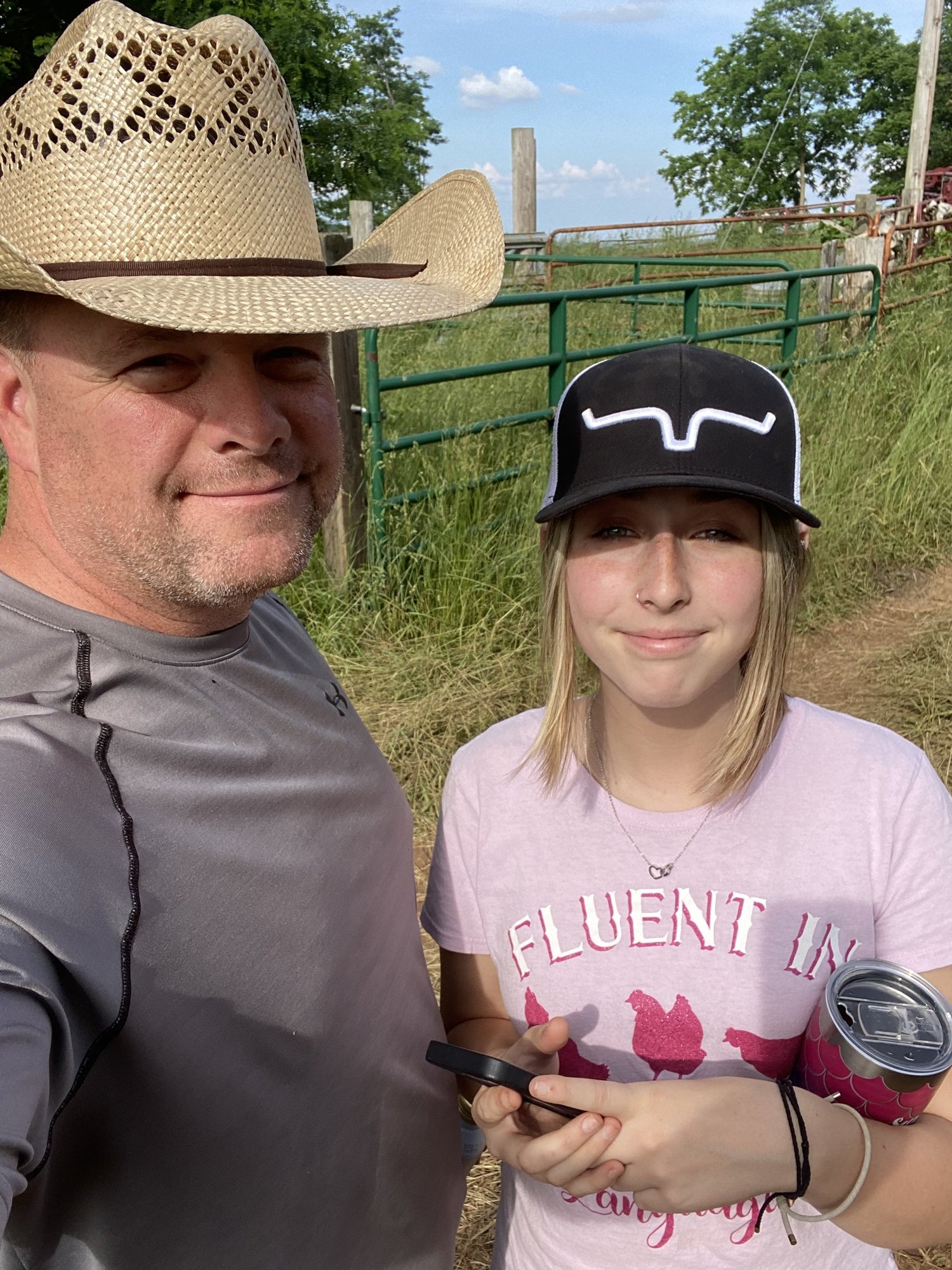 A smiling man in a straw cowboy hat poses for a selfie with a young girl wearing a black cap and a pink shirt that reads "Fluent in Chicken." They are outdoors near a green gate, surrounded by tall grass and trees, enjoying a sunny day. The girl is holding a phone and a drink can.