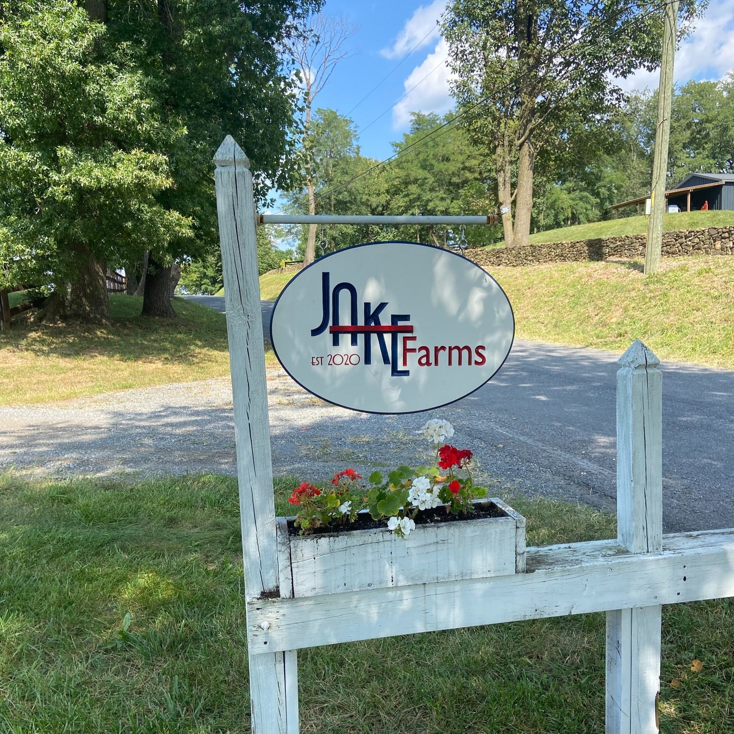 Sign for Jake Farms established in 2020, featuring a flower planter with red and white flowers, located along a gravel road surrounded by trees.