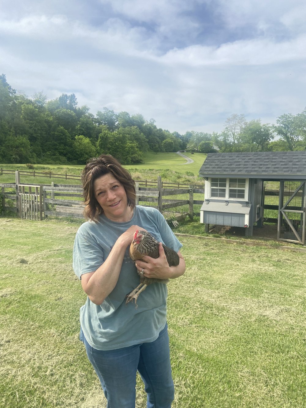 Woman holding a chicken in a grassy field near a chicken coop, with trees and a winding road in the background, showcasing backyard farming and animal husbandry.
